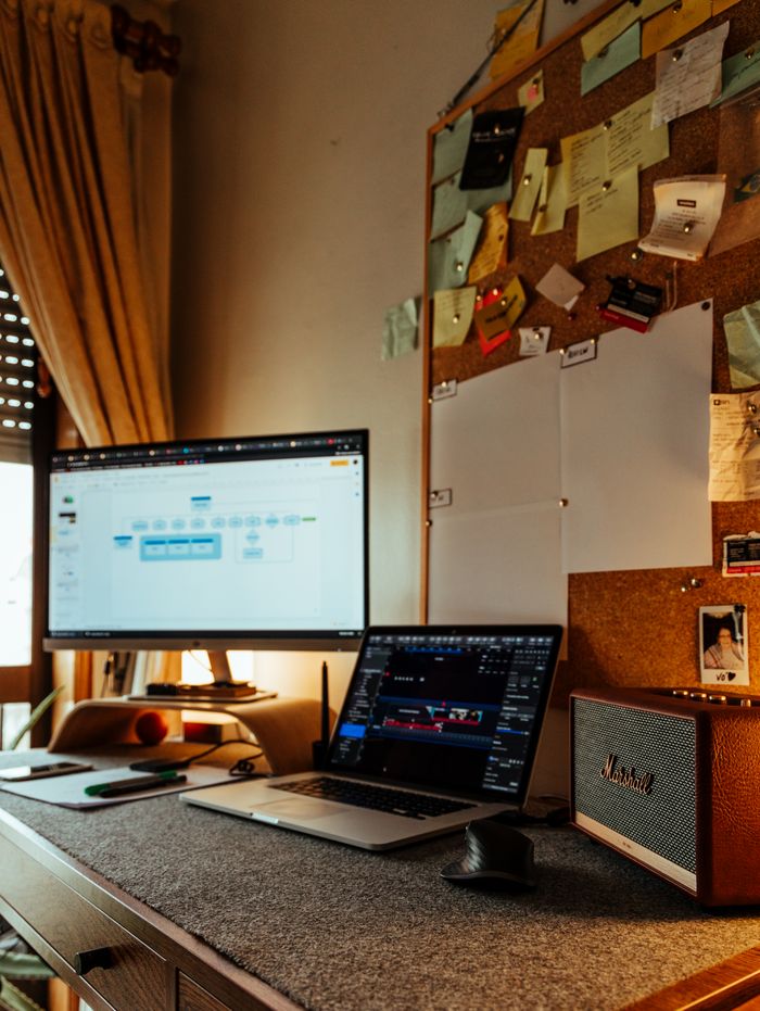 A cozy home office setup with a laptop, monitor, and speaker on a table beside a bulletin board.